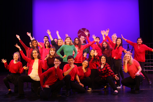 Image Description: A group of student singers dressed in festive holiday colors (red & green) strike a pose on the PAC stage.