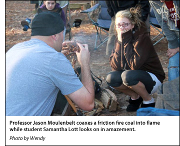 Professor Jason Moulenbelt coaxes a friction fire coal into flame while student Samantha Lott looks on in amazement