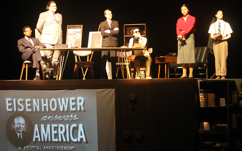 Image Description: A still from "Daisy," Spring 2024. A group of men in suits and one woman look out into the audience. Below them on a giant screen, you can see an old add for Dwight Eisenhower's election..