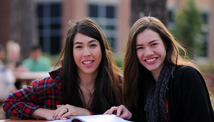 Two female students, smiling at the camera, with a book laying open in front of them
