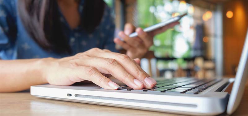 A pair of hands; on on a laptop keyboard, the other holding a mobile device.