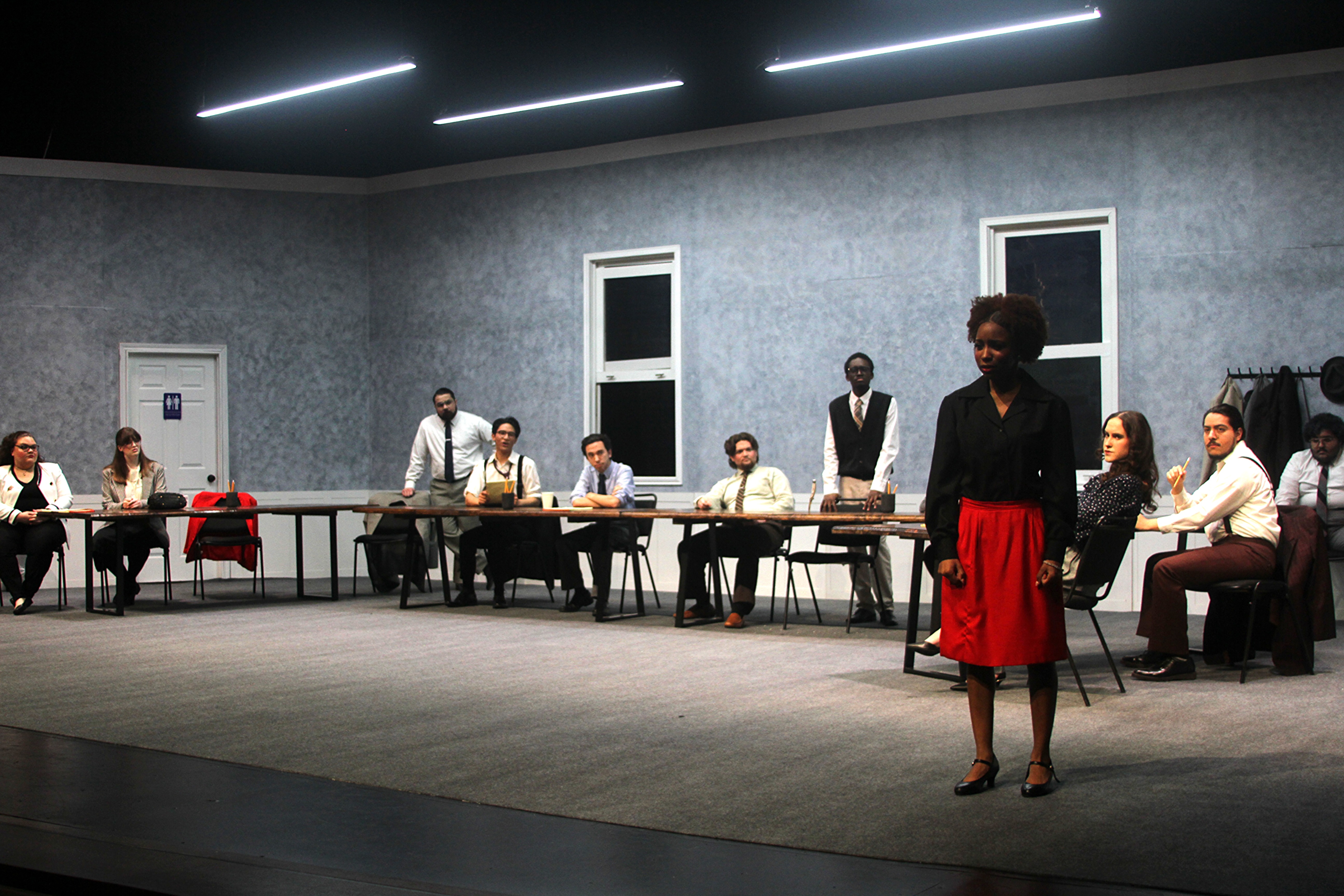 Image Description: A still from "Twelve Angry Jurors," Spring 2026. A group of people in suits look at a lone woman in a starkly lit room.