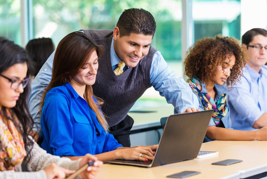 professor looking over the shoulder of a student while she works on a laptop with other students around her