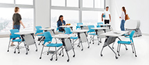 photo of a room with big windows and several flip-top active tables, essentially, a combo of a small desk and chair on casters. a few students are also shown in the room