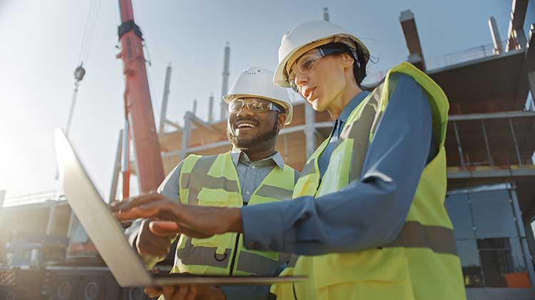 Two workers in front of construction site looking at a laptop screen