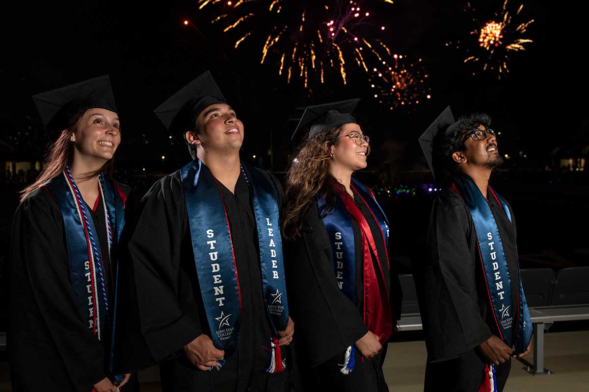 4 students in cap and gown with fireworks in the background.