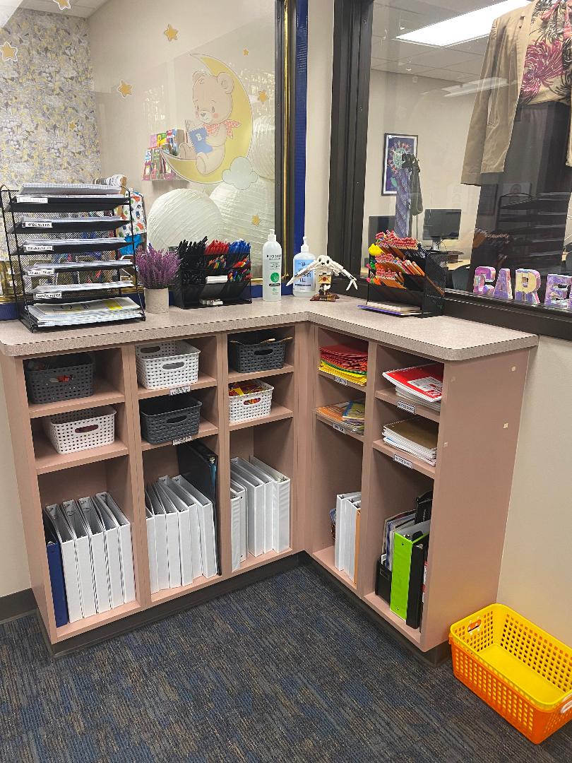 A photo of the school supply corner in the main room of the SRC. A  corner shelf holds many kinds of paper, writing utensils, notebooks, binders, and other school supplies.
