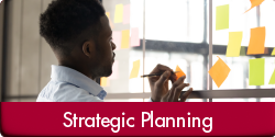Strategic Planning (Photo of a person writing on a sticky note, adding it to several others affixed to glass in an office space. "Strategic Planning" appears in a red bar along the bottom of the image.)