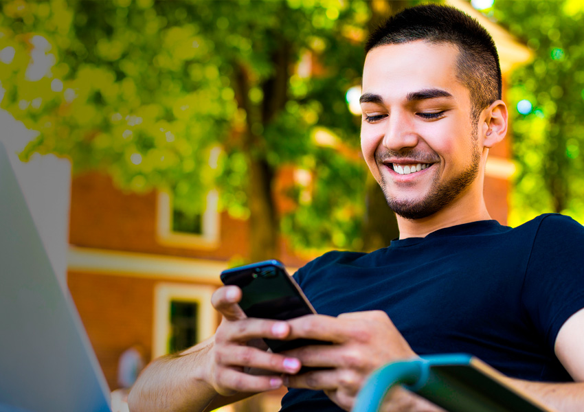 Smiling Male Student Sitting Outdoors and Reading Mobile Phone